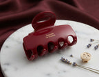 A glossy deep red hair claw clip with gold lettering reading 'Barette La France', displayed on a white marble tray with lavender sprigs and a gold hair pin against a draped burgundy fabric background.
