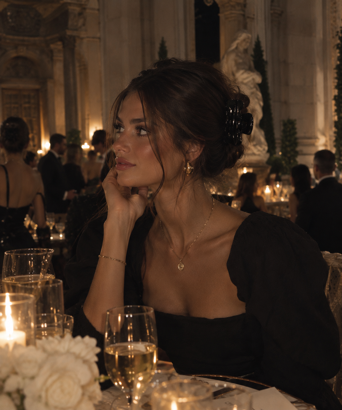 A woman in a black off-the-shoulder dress sits at a candlelit dinner table in an elegant indoor venue, wearing gold jewelry including a necklace and earrings. The elegant black hair clip is keeping the hairs together